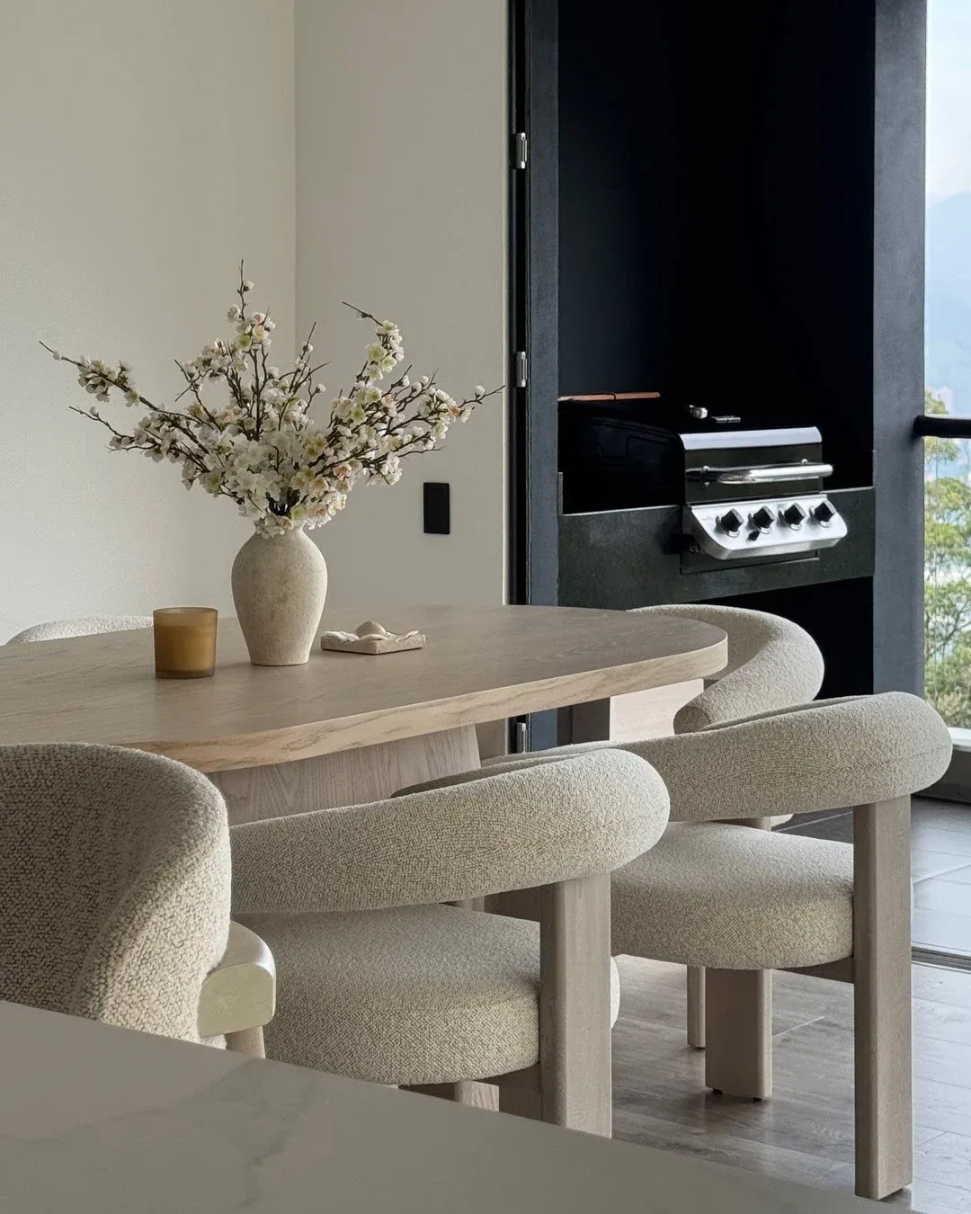 Mesa de comedor de madera con flores, con vista a un balcón con zona de barbacoa.