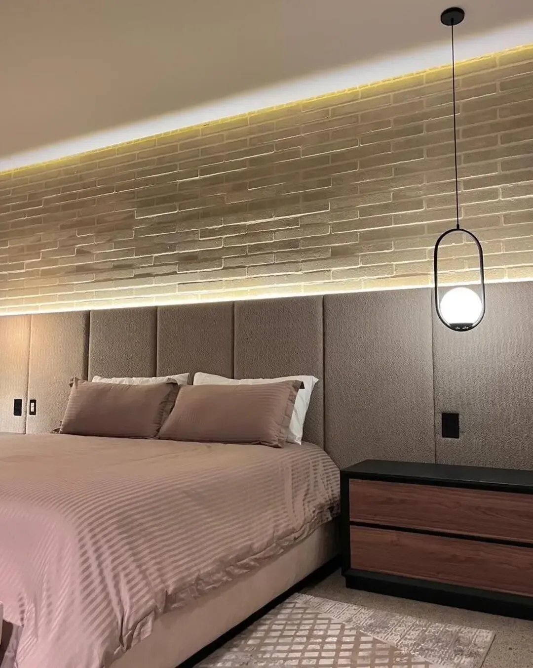 Dormitorio industrial con cabecero tapizado, muro en ladrillo e iluminación cálida envolvente.