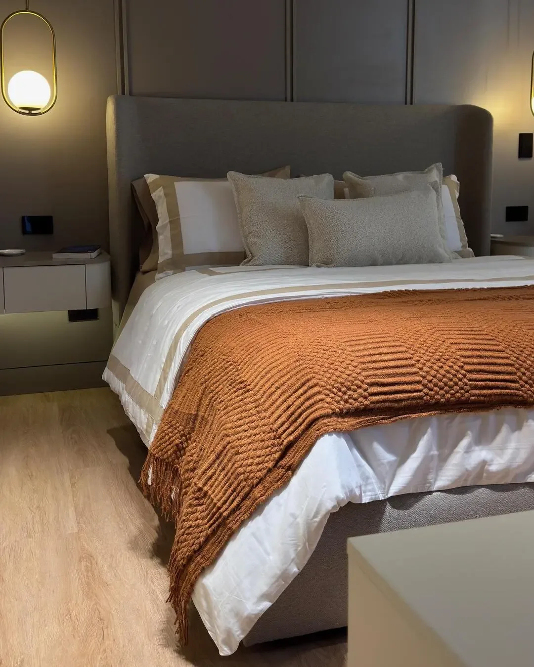 Dormitorio cálido con cabecero tapizado, textiles neutros y luz suave de acento.