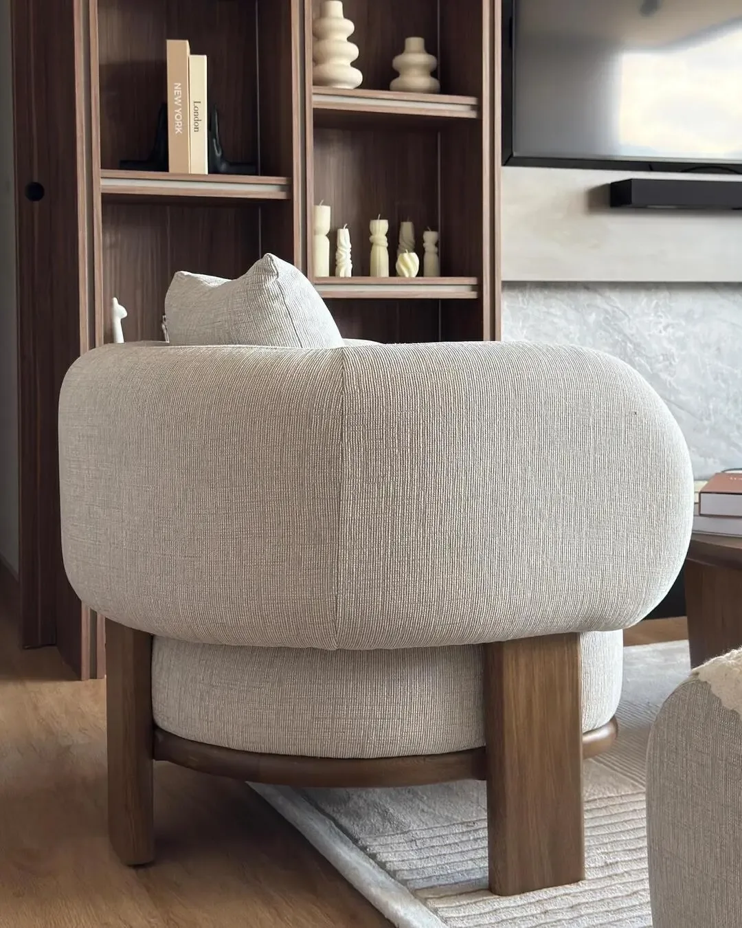Silla auxiliar envolvente con madera natural, tapizado claro y proporciones ergonómicas.