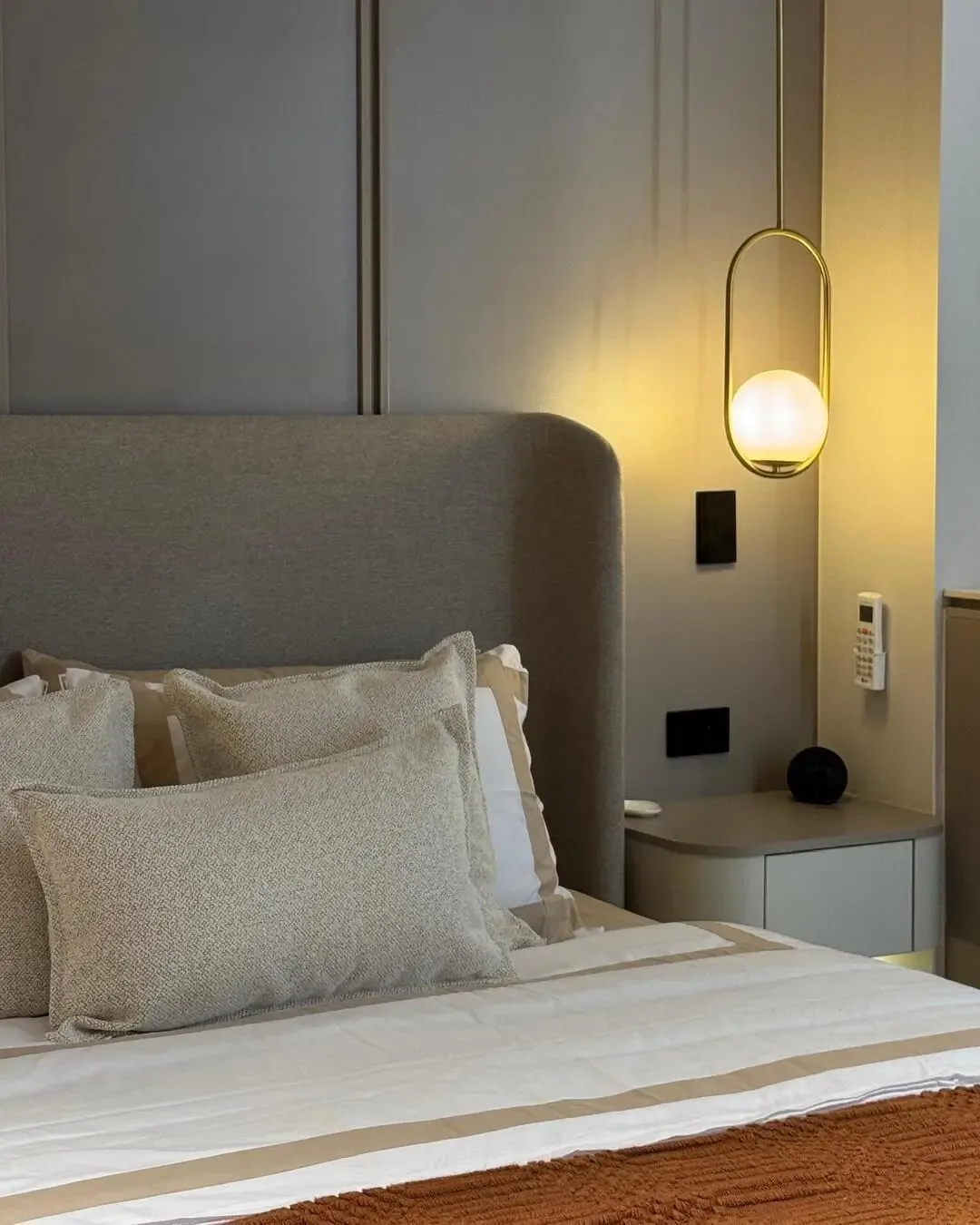 Detalle de cama tapizada con lámpara suspendida, cojines texturizados y tonos cálidos.