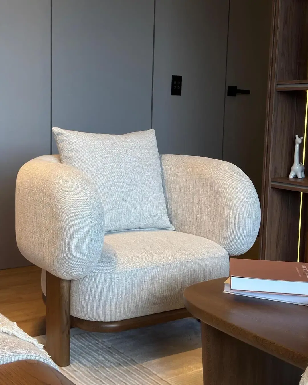 Silla tapizada con brazos redondeados, madera natural y carácter cálido contemporáneo.