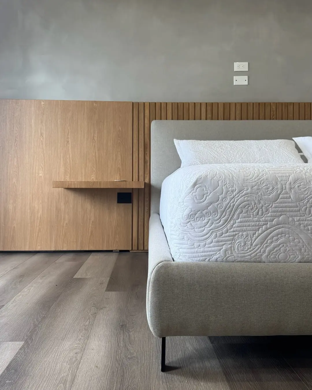 Detalle de una mesa de noche redonda en madera junto a una cama con cabecero de paneles grises.