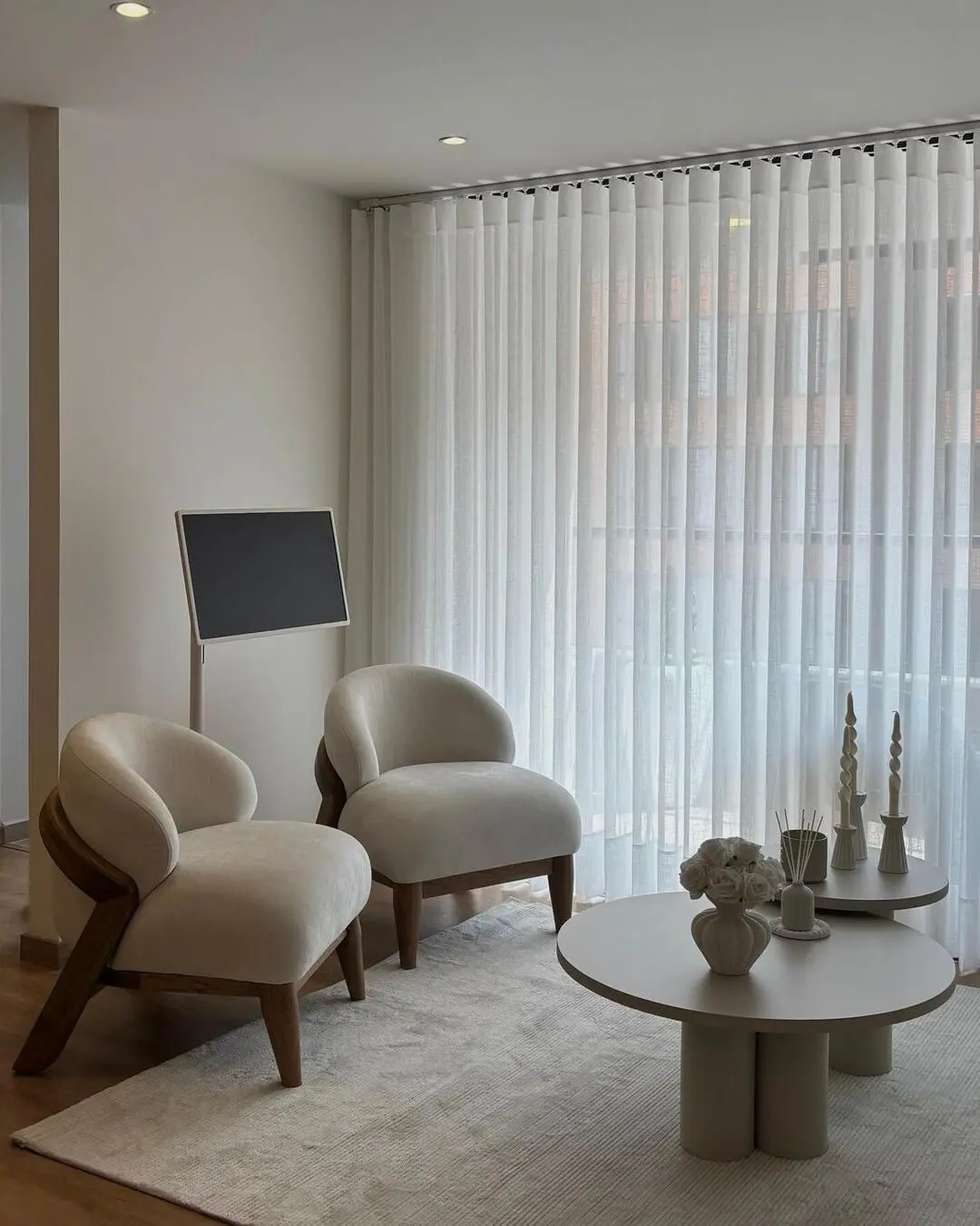 Vista de una sala de estar con dos poltronas blancas, mesas de centro y cortinas translúcidas de fon